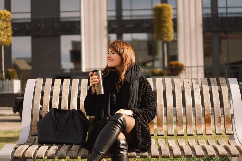A woman sits on a bench in the city and drinks coffee and thermal cups.