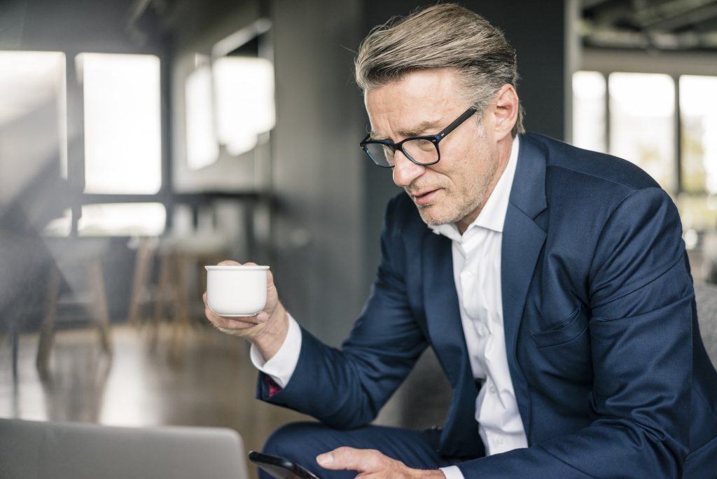 Mature businessman with cup of coffee and laptop using cell phone